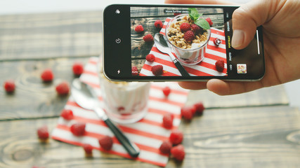 Hand of anonymous person holding modern smartphone and making lovely photo of delicious fruit dessert and spoon on wooden table