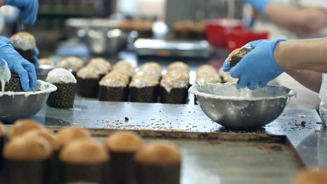 Tracking shot of hands of unrecognizable confectioners in gloves covering Easter bread in frosting and decorating it at factory