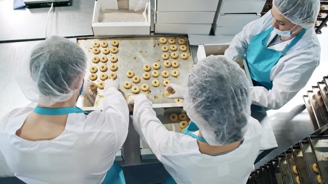 High angle view of three confectionery workers in workwear, bouffant mob caps, aprons and face masks packing cookies at factory before sending to shops
