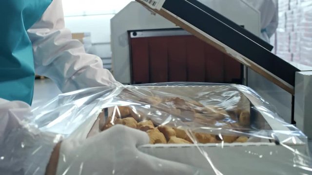 Tilt down shot of woman wrapping box with fresh cookies in transparent foil and sealing it using factory machine