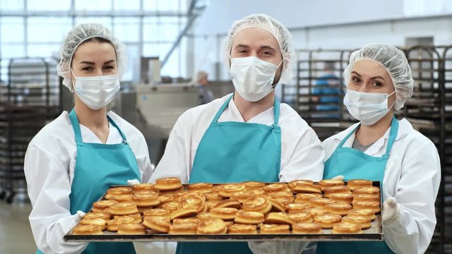 Portrait of three friendly factory workers in bouffant mob caps, aprons and face masks holding baking tray with delicious freshly baked cookies and looking at camera