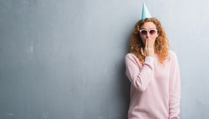 Young redhead woman over grey grunge wall wearing birthday cap cover mouth with hand shocked with shame for mistake, expression of fear, scared in silence, secret concept