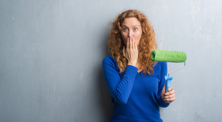 Young redhead woman over grey grunge wall holding painting roller cover mouth with hand shocked with shame for mistake, expression of fear, scared in silence, secret concept