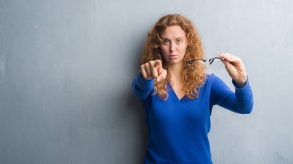 Fototapeta premium Young redhead woman over grey grunge wall holding optical glasses pointing with finger to the camera and to you, hand sign, positive and confident gesture from the front