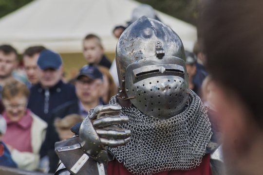 rycerz zakuty w zbroję z metalową rękawicą, za rycerzem rozmyty tłum widz&oacute;w, przed rycerzem rozmyta sylwetka głowy