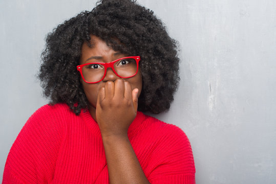 Young african american plus size woman over grey grunge wall wearing winter sweater looking stressed and nervous with hands on mouth biting nails. Anxiety problem.