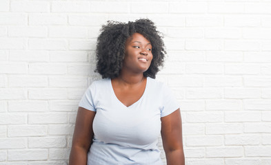 Young african american plus size woman over white brick wall looking away to side with smile on face, natural expression. Laughing confident.