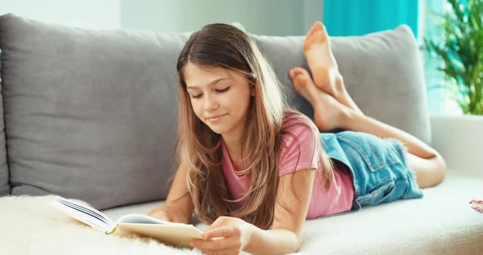 Teenage girl reading book, smiling, lying on the sofa indoors
