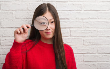 Young Chinese woman over brick wall looking through magnifying glass with a confident expression on smart face thinking serious