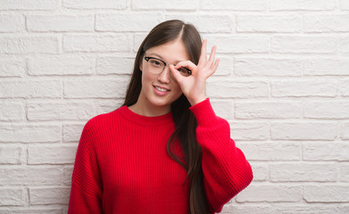 Young Chinese woman over brick wall with happy face smiling doing ok sign with hand on eye looking through fingers