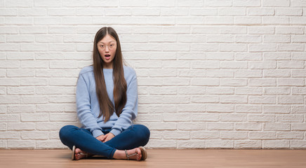 Young Chinese woman sitting on the floor over brick wall afraid and shocked with surprise...