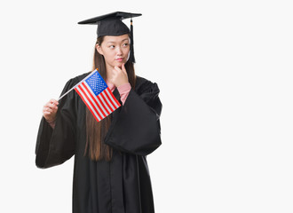 Young Chinese woman wearing graduate uniform holding United states of america flag serious face thinking about question, very confused idea