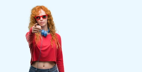 Young redhead woman wearing headphones pointing with finger to the camera and to you, hand sign, positive and confident gesture from the front