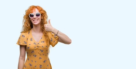 Young redhead woman wearing sumer outfit and sunglasses happy with big smile doing ok sign, thumb up with fingers, excellent sign