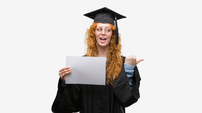 Young redhead woman wearing graduate uniform holding degree pointing and showing with thumb up to the side with happy face smiling