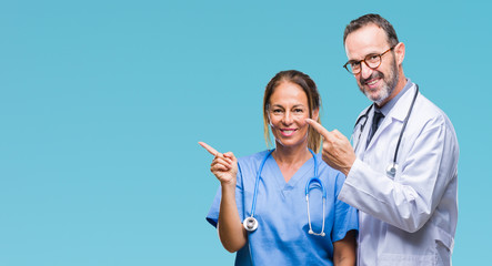 Middle age hispanic doctors partners couple wearing medical uniform over isolated background smiling and looking at the camera pointing with two hands and fingers to the side.
