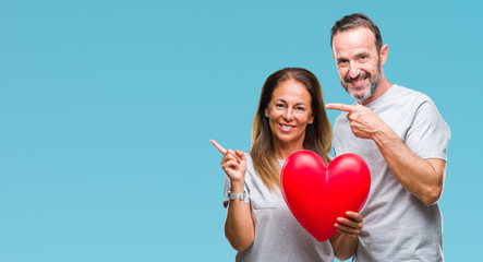 Middle age hispanic casual couple in love holding red heart over isolated background very happy pointing with hand and finger to the side