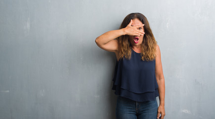 Middle age hispanic woman standing over grey grunge wall peeking in shock covering face and eyes with hand, looking through fingers with embarrassed expression.