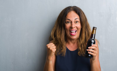 Middle age hispanic woman standing over grey grunge wall holding beer bottle screaming proud and celebrating victory and success very excited, cheering emotion