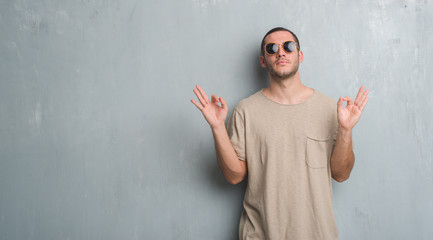 Young caucasian man over grey grunge wall wearing sunglasses relax and smiling with eyes closed doing meditation gesture with fingers. Yoga concept.
