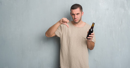 Young caucasian man over grey grunge wall holding bottle beer with angry face, negative sign...
