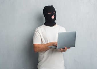 Young caucasian hacker man over grey grunge wall doing cyber attack using laptop with a happy face...