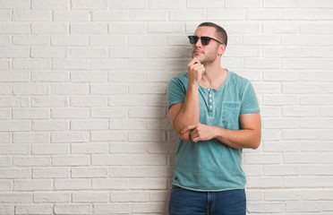 Young caucasian man standing over white brick wall wearing sunglasses serious face thinking about question, very confused idea