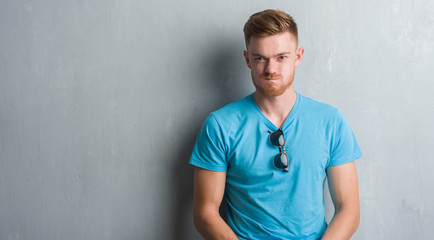 Young redhead man over grey grunge wall wearing casual outfit puffing cheeks with funny face. Mouth inflated with air, crazy expression.