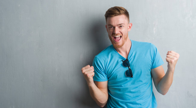 Young redhead man over grey grunge wall wearing casual outfit very happy and excited doing winner gesture with arms raised, smiling and screaming for success. Celebration concept.