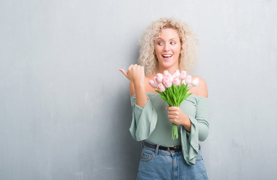 Young blonde woman over grunge grey background holding flowers pointing and showing with thumb up to the side with happy face smiling