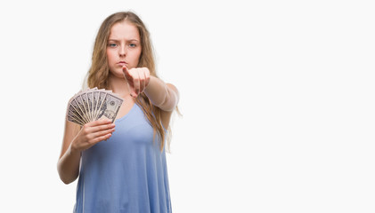 Obraz premium Young blonde woman holding dollars pointing with finger to the camera and to you, hand sign, positive and confident gesture from the front