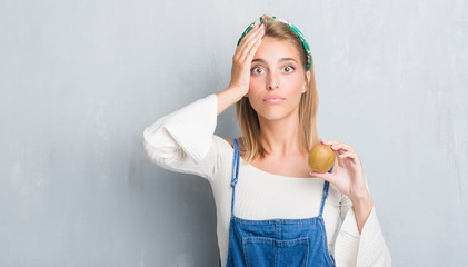 Beautiful young woman over grunge grey wall holding fresh kiwi stressed with hand on head, shocked with shame and surprise face, angry and frustrated. Fear and upset for mistake.