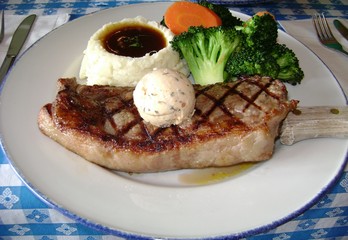 A huge slab of medium rare beef steak in a plate, served with a butter ball, mashed potatoes and vegetables.