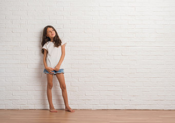 Young hispanic kid stading over white brick wall looking away to side with smile on face, natural...