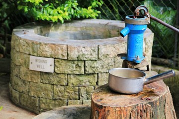 An old outdoors water pump with a pan to catch the water beside a round cement well 