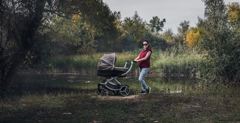Young stylish beautiful mother walks with a child in a stroller in the woods along the river, dressed in jeans, a red vest and glasses. Panorama, flash lightс