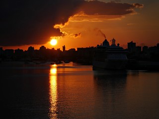 Fototapeta premium A big round sun sets over the port of Old San Juan in Puerto Rico, casting golden fiery reflections in the water. 