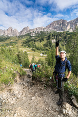 young male climber points up  at a Via Ferrata in the Dolomites and many people stand behind him