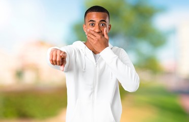 Dark-skinned young man with white sweatshirt pointing with finger at someone and laughing a lot while covering mouth in a park