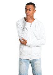 Dark-skinned young man with white sweatshirt standing and looking to the side with the hand on the chin on isolated white background