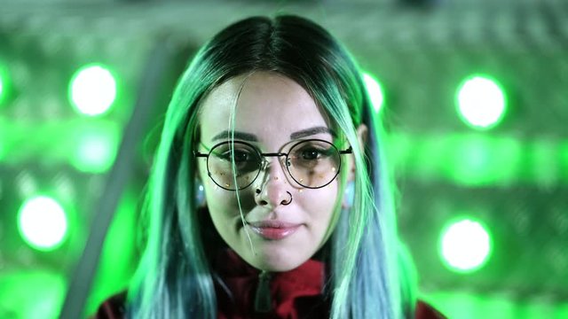 Hipster girl with blue dyed hair and golden sequins as freckles. Woman with nose piercing, transparent glasses, ears tunnels, unusual hairstyle stands in amusement night park