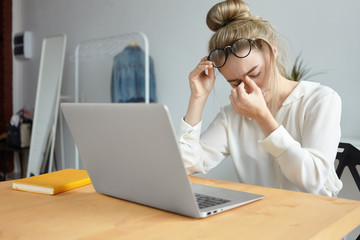 Modern technology, job and people concept. Portrait of tired young female employee with hair bun taking off eyeglasses and massaging her nose bridge, feeling stressed because of lot of work - obrazy, fototapety, plakaty