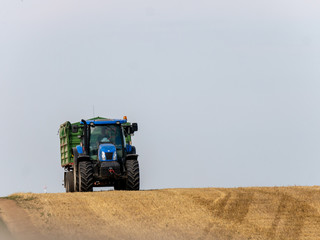 Obraz premium Blue tractor with trailer on the harvest field. Summer field and blue tractor with trailer. Harvest field, blue sky and tractor.