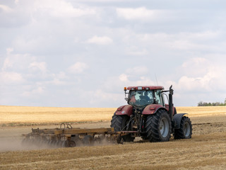 Obraz premium Red tractor with plow on harvest field. Summer field and red tractor. Tractor on harvest field. Lowing after harvest. Plowing on the field.