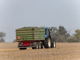 Blue tractor with trailer on the harvest field. Summer field and blue tractor with trailer. Harvest field, blue sky and tractor.