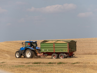 Obraz premium Blue tractor with trailer on the harvest field. Summer field and blue tractor with trailer. Harvest field, blue sky and tractor.