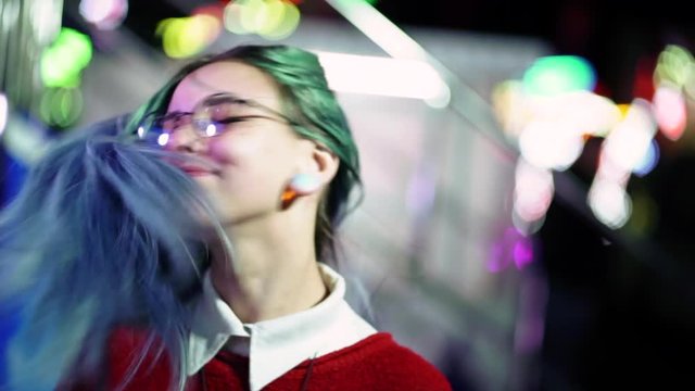 Hipster girl waving head in different directions, playing with dyed blue hair. Woman with nose piercing, transparent glasses, ears tunnels, unusual hairstyle in amusement park.