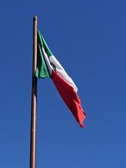 Flag of Mexico flying halfway unfurled in the air against a backdrop of blue skies