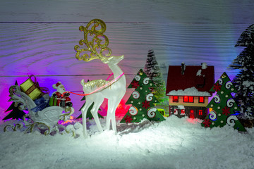 Christmas composition. Santa Claus in a sleigh with gifts, a fairy-tale white deer, a toy small house with decorative lighting a midst snow-covered firs.