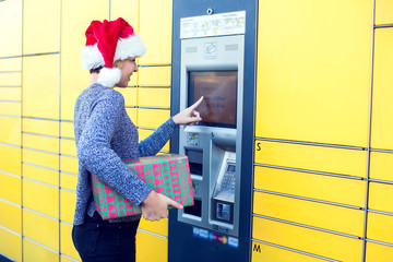 Woman with santa hat client using automated self service post terminal machine or locker to deposit the parcel for storage